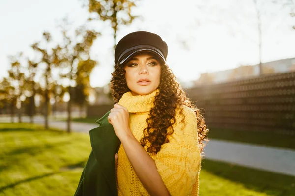 In het park poseert het meisje voor een foto op haar gezicht een vastberaden uitdrukking op één schouder een jas aan die contrasteert met een felgele trui — Stockfoto