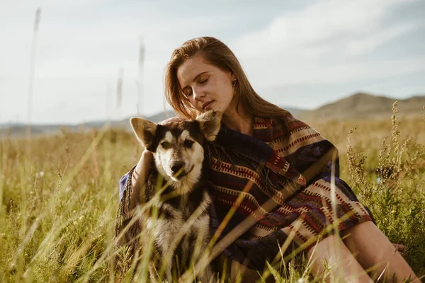 In the steppe against the backdrop of hills on the grass sit a girl and her dog. the girl is wrapped in a shawl and covered her eyes cuddling up to an eared friend — Stock Photo, Image