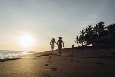 Güneşin arka planında, kumsalda okyanusta oturan genç adam ve kız el ele tutuşurken mesafeye koşun.