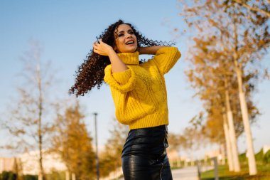 Parlak sarı kazaklı bir kız, sarı sonbahar ağaçlarının arka planında rüzgarda dalgalı kıvırcık saçlarını büker.