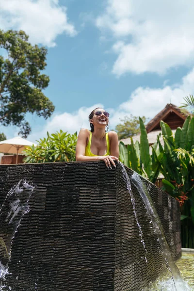 A mulher europeia descansando em óculos de sol e um maiô amarelo inclinado na borda da piscina e olhando para fora do lado olhando para a distância sorrindo afetuosamente — Fotografia de Stock