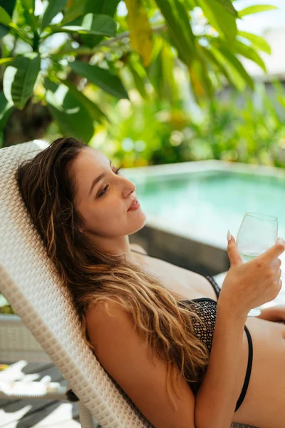Το κορίτσι αναπαύεται στην πισίνα στο φόντο των φοινίκων και κρατά ένα ποτήρι κρύο ποτό στο χέρι της — Φωτογραφία Αρχείου
