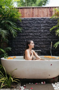 Çiçek tomurcuklarıyla banyodaki spa tedavileri taş bir duvarın ve palmiye yapraklarının arkasında uzun ıslak su saçı olan bir kızı oturtuyor.