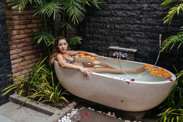 Спа-ванна с бутонами желтых цветов на фоне каменных стен и пальм. В ванной сидит красивая девушка с длинными волосами в купальнике — стоковое фото