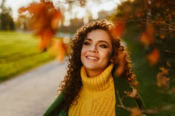 Los preciosos dientes fueron expuestos en la sonrisa de una hermosa morena rizada de pie junto a un árbol en un parque de otoño — Foto de Stock