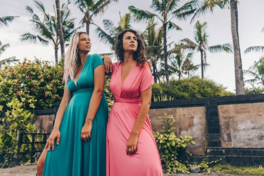 Parlak uzun elbiseli esmer ve sarışın, palmiye ağaçları ve villanın çitlerine karşı poz veriyor.