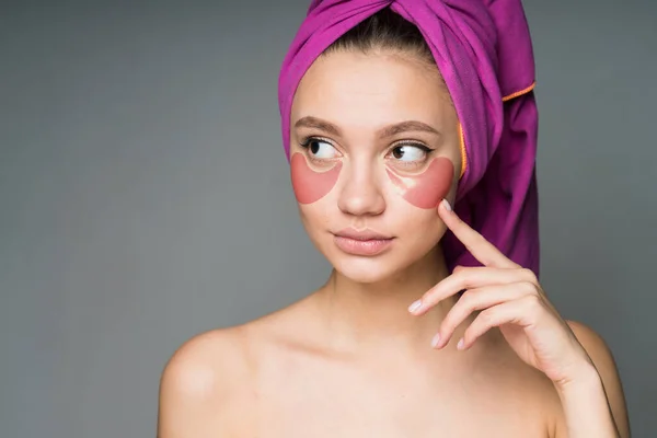 Girl with a towel wrapped around her head, bare shoulders makes herself a spa procedure by sticking patches under her eyes — Stock Photo, Image