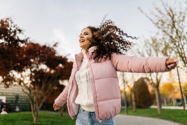 Kollarını kenarlara uzatarak parkta bir gülümseme içinde ağzını açarak pembe ceketli ve kot pantolonlu kanat çırpan bir kız koşar biriyle buluşmak için.