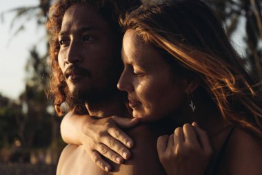 Doğu Asyalı bir adam mesafeye bakar ve arkasında bir kız durur ve onu boynundan sarar.