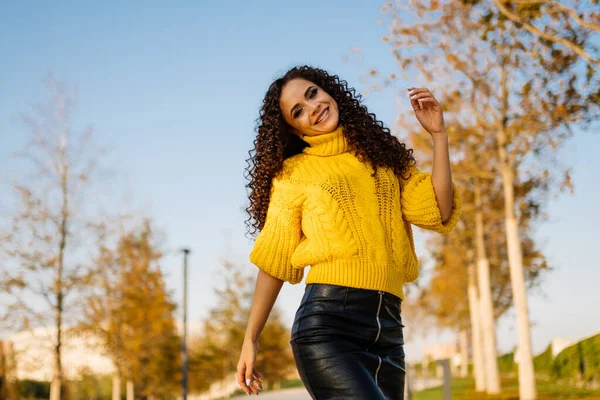 A very cute smile on the face of a curly beauty in a bright yellow sweater posing in an autumn park — Stock Photo, Image