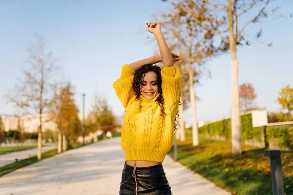 Uma menina em uma camisola amarela brilhante dança no caminho do parque de outono levantando as mãos e sorrindo flertando — Fotografia de Stock