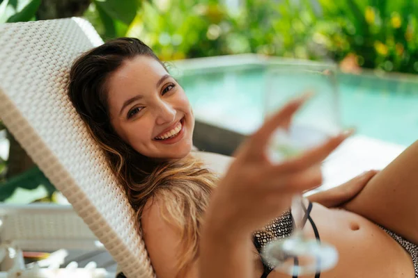 У бассейна на шизонг с бокалом в руке лежит очаровательно улыбающаяся девушка в купальнике — стоковое фото