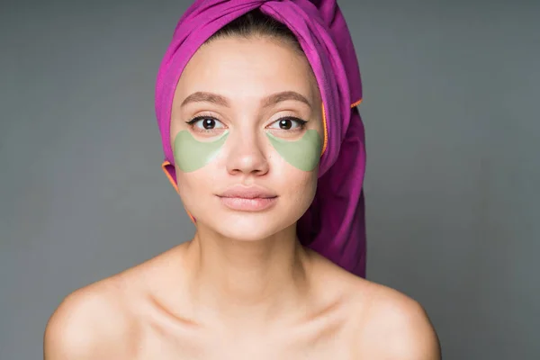 Menina com ombros nus e toalha na cabeça faz procedimento spa, colocando manchas sob os olhos — Fotografia de Stock