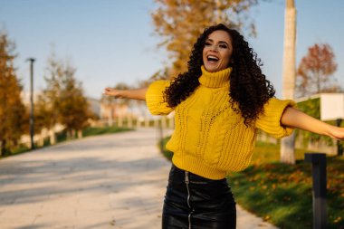 Sonbahar parkında parlak bir süveter içinde kollarını kıvırcık esmerin kenarına uzatarak sevinçle çığlıklar atıyor.