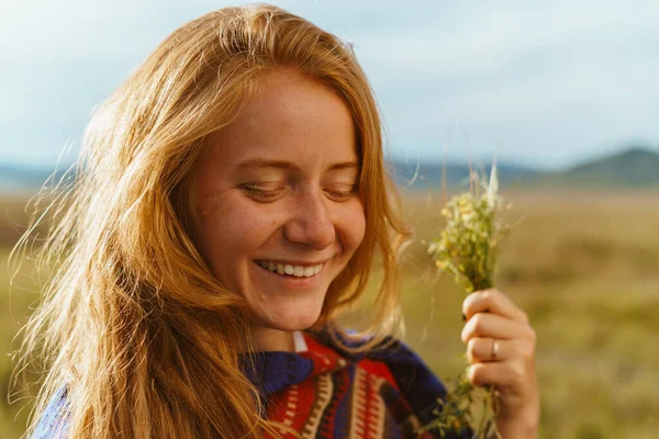 Beleza natural de uma menina ruiva com sardas um sorriso suave segurando um buquê de flores silvestres no fundo da estepe e colinas. close-up — Fotografia de Stock