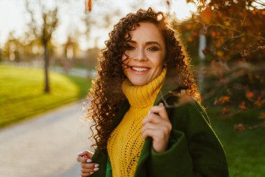 Yakın plan. Kıvırcık saçlı, güzel, beyaz dişli bir gülüşü olan esmer, sonbahar parkında poz veriyor.