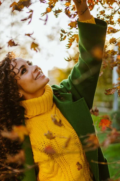 Uma menina em um suéter amarelo brilhante do outono toca as folhas amareladas na árvore e sorri suavemente com um sorriso bonito — Fotografia de Stock