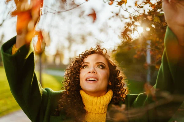 Una niña en el parque de otoño en los rayos del sol toca las manos de las hojas de otoño en las ramas de un árbol — Foto de Stock