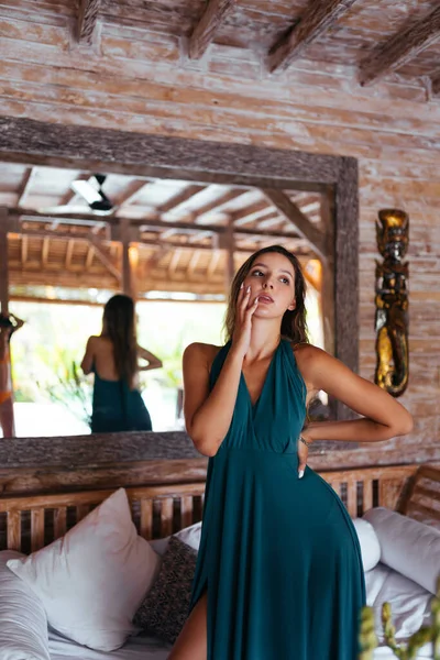 A menina no bangalô fica perto do sofá em um vestido longo e se toca com a mão sobre a bochecha levantando os olhos — Fotografia de Stock