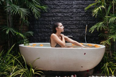 Banyoda, küçük palmiye ağaçlarının arasında taş bir duvarın arkasında bir kız oturuyor.