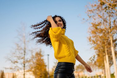 Mavi gökyüzüne karşı parlak sarı bir kazak ve kıvırcık saçlı neşeli gülümseyen bir kızla dans eden ağaçların sarı yaprakları.