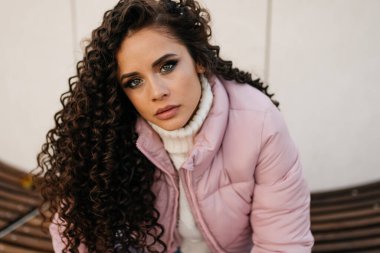 Yakın plan. Koyu renk saçlı kıvırcık saçlı, yeşil gözlü ve makyajlı bakışlı. Sıcak giysiler içinde bir şeyler çalışırken dikkat çeken biri.