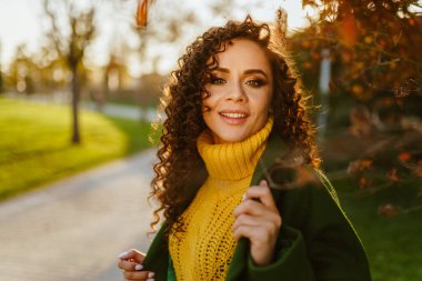 Güneşli bir sonbahar gününde, bir sonbahar parkında uzun kıvırcık saçlı bir kız gururla gülümseyerek pürüzsüz beyaz dişlerini gösteriyor.
