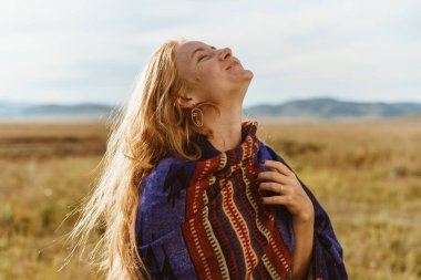 Kırmızı saçlı, etno pelerinli, gülen bir kızın kafasını tepelerin arkasındaki bozkıra fırlatıyor.