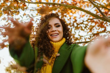 Neşeli bir şekilde ellerini öne çekiyor ve güzel kıvırcık esmer gülüyor sonbahar yapraklarının arka planına doğru.