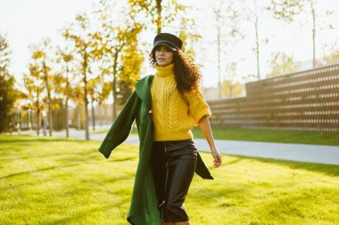 Sarı, kalın kazaklı ve şapkalı bir kız tek omuzlu bir ceketle çimenlikte hızla yürüyor.