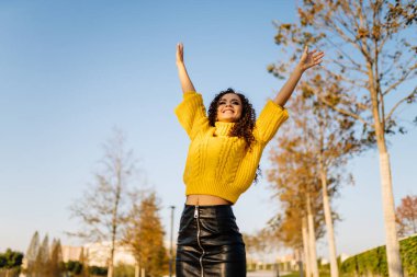 Mavi gökyüzüne ve sonbahar parkına karşı parlak bir kazak ve deri bir etek giyen mutlu ve mutlu bir şekilde ellerini kaldıran bir kız..