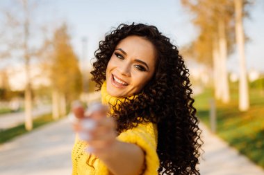 Siyah, kıvırcık saçlı, gülen bir kız, elini kameraya çeker sonbahar ağaçlarıyla dolu bir arka sokağın arka planına.