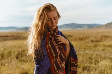 Buğday rengi saçları olan bir kız başını eğdi ve gözlerini eğdi etno bir pelerine sarılmış bir şekilde bozkırda duran tepelerin arkasında duruyordu.