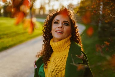 Sarı-yeşil sonbahar parkının arka planında parlak sarı bir süveter ve yeşil bir ceket içindeki koyu renk saçlı güzelin coşkulu bakışları