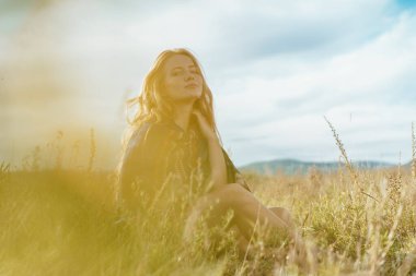Gökyüzüne ve bozkır çimlerine karşı, saçları omuzlarına yayılmış ve boynuna elleriyle dokunan bir kadın.