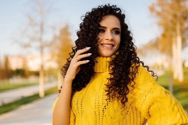 Parlak sarı bir süveter içinde kıvırcık saçlı, geniş gülüşlü güzel bir kadın koyu saçlarını sonbahar parkının arka planına doğru düzeltiyor.