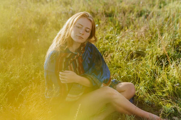 En la hierba del prado se sienta una chica envuelta en una capa etno exponer piernas delgadas su pelo de trigo extendido sobre sus hombros — Foto de Stock