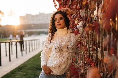 Kırmızı yapraklarla çevrili duvarda dirseğine yaslanmış sıradan giyinmiş şık bir kız duruyor.