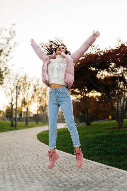 Kalın elbiseli bir kız, saçları süzülürken kollarını havaya kaldırarak parkta yürüyüş yolunda zıplıyor.