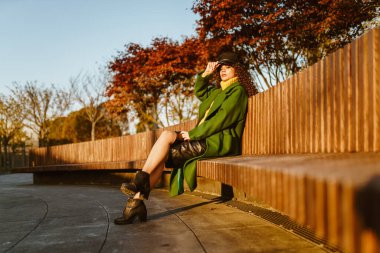 Parktaki uzun ahşap bankta mor sonbahar ağaçlarının arka planında şık giyinmiş sıcak sonbahar kıyafetleri içinde bir kız oturuyor.