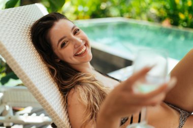 Çekici bir şekilde gülümseyen bir kız kameraya bir bardak uzatır ve bir havuz ve palmiye ağaçlarına yaslanır. yakın plan