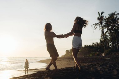 Bir adam ve bir kadın el ele tutuşarak kumun üzerinde dönüyor okyanus ve palmiye ağaçlarına karşı.