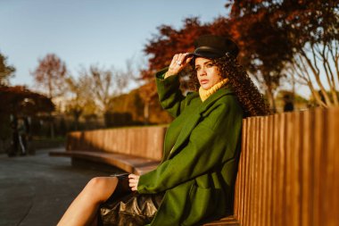 Parkta bankta sonbahar elbiseleriyle oturan kız ayağını bacağına atıyor.