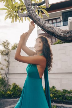 Mavi elbiseli neşeli bir kız beyaz dişli bir gülümseme ile çitin arkasındaki otelin arka planındaki ağacın altında dans ediyor.