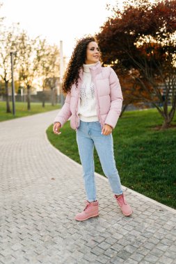 Parktaki yürüyüş yolunda, sonbahar kıyafetleri içinde rüzgarı dalgalı saçlar ve gülümseyen bir kız duruyor.