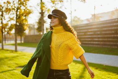 Parkın yeşil çimlerinde bir kız koşar bir omuzuna sonbahar montu atar