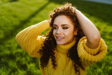 Parlak sarı kazaklı yeşil çimen kızı yakından çek. Kıvırcık saçlarını elleriyle kaldırıyor.