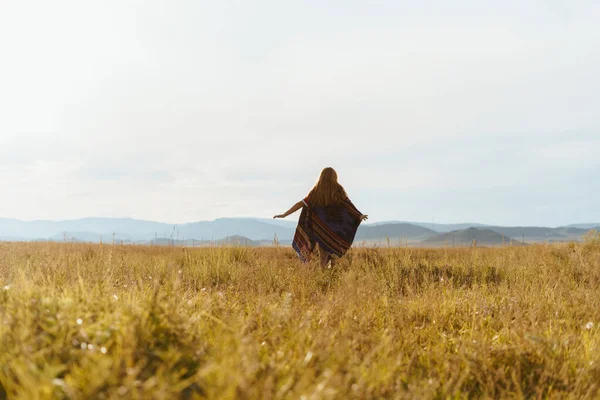 Corre hacia la distancia extendiendo sus brazos en una capa etnográfica sobre los hombros de una chica en el prado de hierba hacia las colinas — Foto de Stock