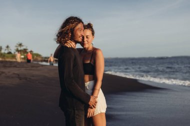 Okyanus sularının ve sahil kenarındaki kumların ardında bir arkadaşa sokulmuş, uzaktan bakan bir adam ve mutlulukla gözlerini kapayan bir kadın.