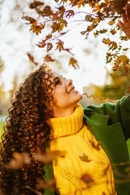 Kıvırcık esmer, kıpkırmızı yaprakların arkasındaki ağacın dallarına kadar kollarını uzattı sonbahar bahçesinde.